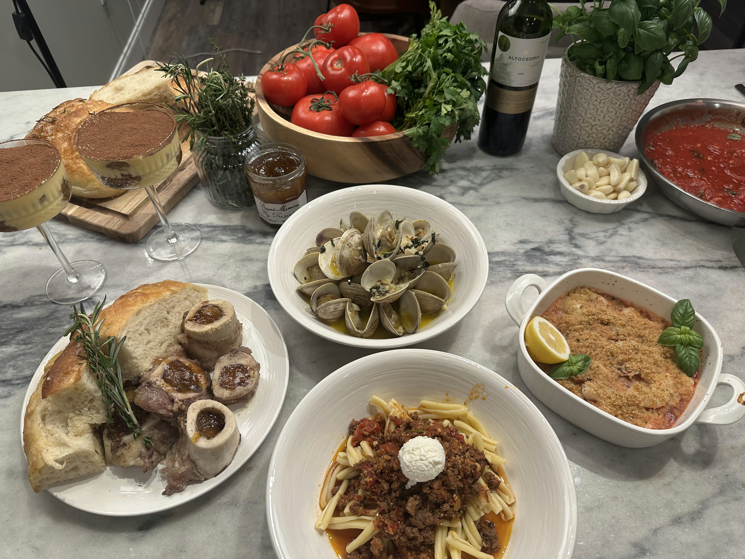 A home-cooked Italian dinner spread with pasta, clams, tomatoes, fresh herbs, bread, and dessert